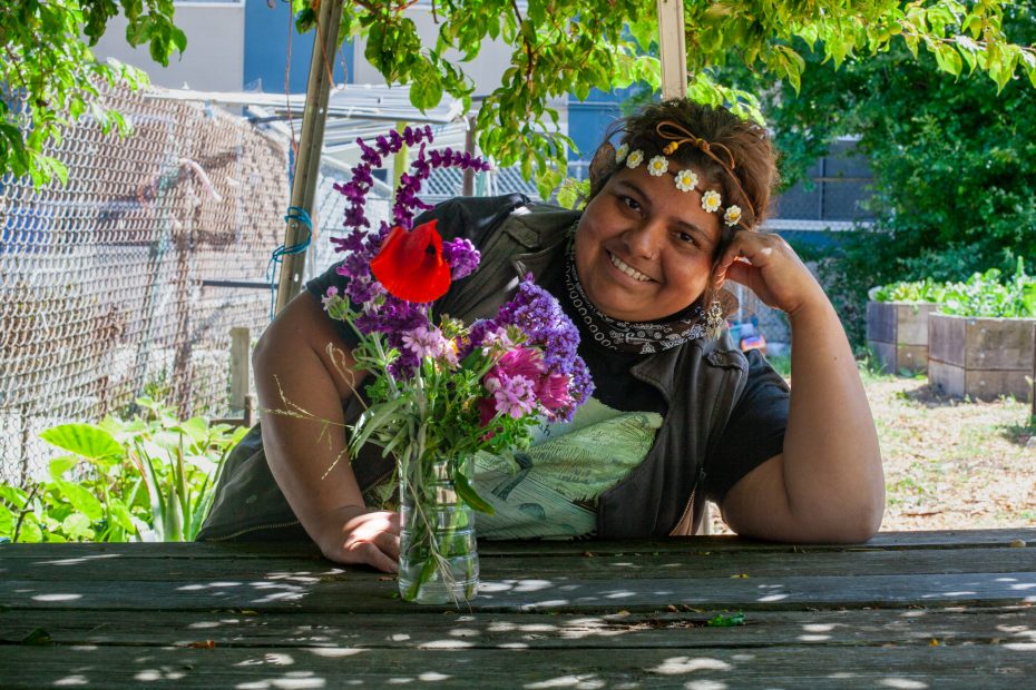 A person wearing a flower crown smiles while leaning on a wooden table with a jar of colorful flowers, under a leafy canopy in a garden setting.