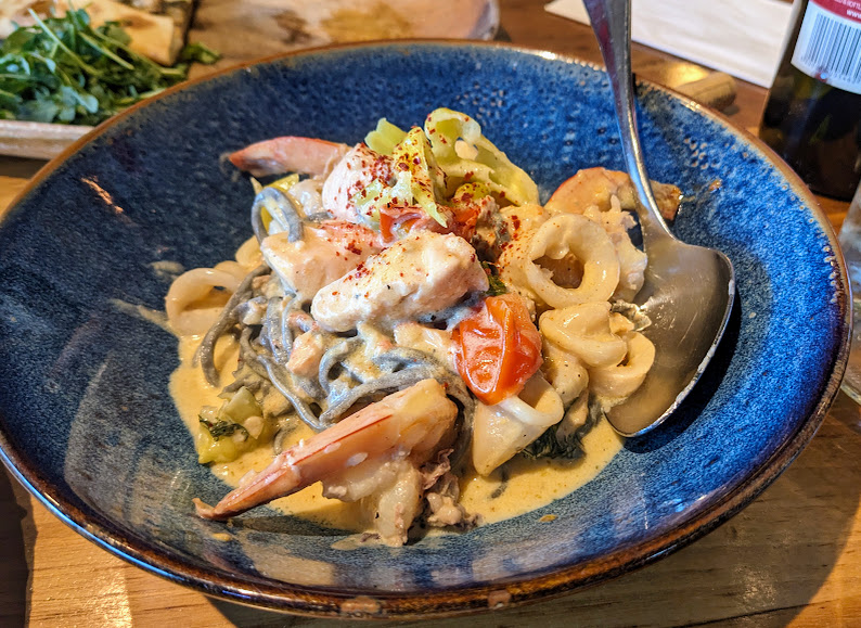 A blue bowl containing creamy seafood pasta with shrimp, calamari, cherry tomatoes, sliced peppers, and a spoon.