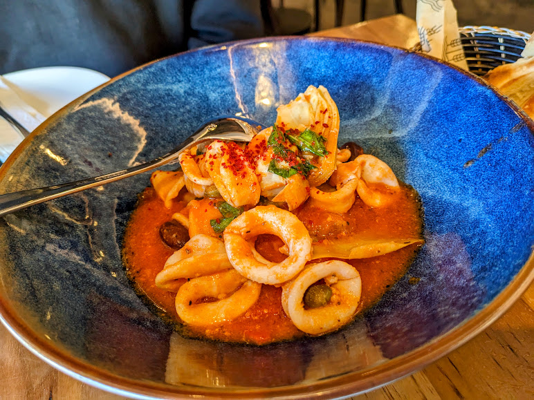 A blue bowl filled with seafood pasta in a red tomato sauce, garnished with herbs and chili flakes, with a fork resting on the side.