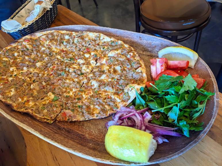 A wooden platter with a baked flatbread topped with minced meat and herbs, served with lemon wedges, sliced tomatoes, parsley, and red onion.