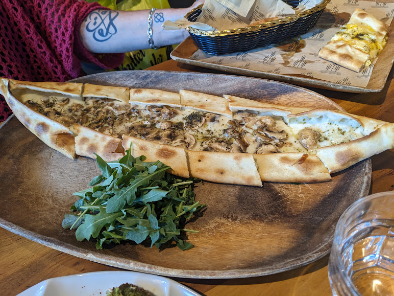 A baked Turkish pide topped with mushrooms and cheese on a wooden platter, served with a side of fresh arugula. Other food items and a tattooed arm are visible in the background.