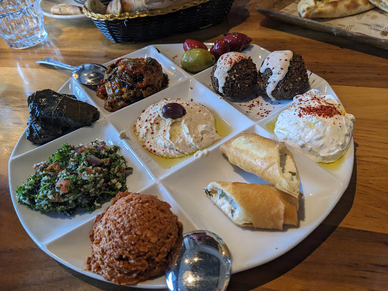 A white plate with various Middle Eastern appetizers including hummus, stuffed grape leaves, tabbouleh, falafel, spanakopita, olives, baba ghanoush, tzatziki, and muhammara.