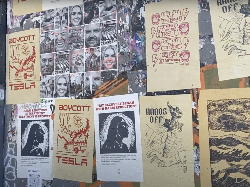 A wall covered with various posters and flyers, including anti-Tesla messages, harm reduction themes, black-and-white portraits, and hand-drawn illustrations.