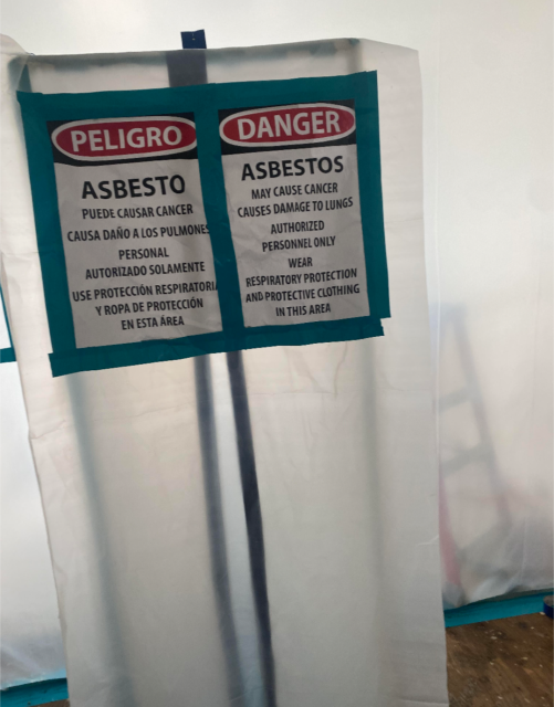 Sign in Spanish and English warning about asbestos, noting cancer and lung risks. Entry restricted to authorized personnel with respiratory and protective gear.