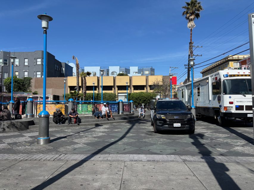 A city plaza with people sitting on benches, a black SUV, and a large white vehicle labeled "Mobile Command" parked nearby under a clear blue sky.
