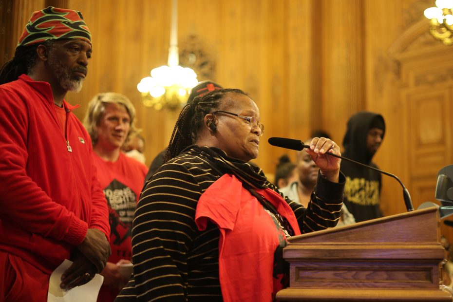 A woman speaks into a microphone at a podium while others stand nearby in a wood-paneled room.