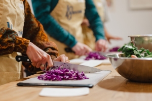 People wearing aprons chop purple cabbage on cutting boards at a kitchen counter, with a bowl of leafy greens nearby.