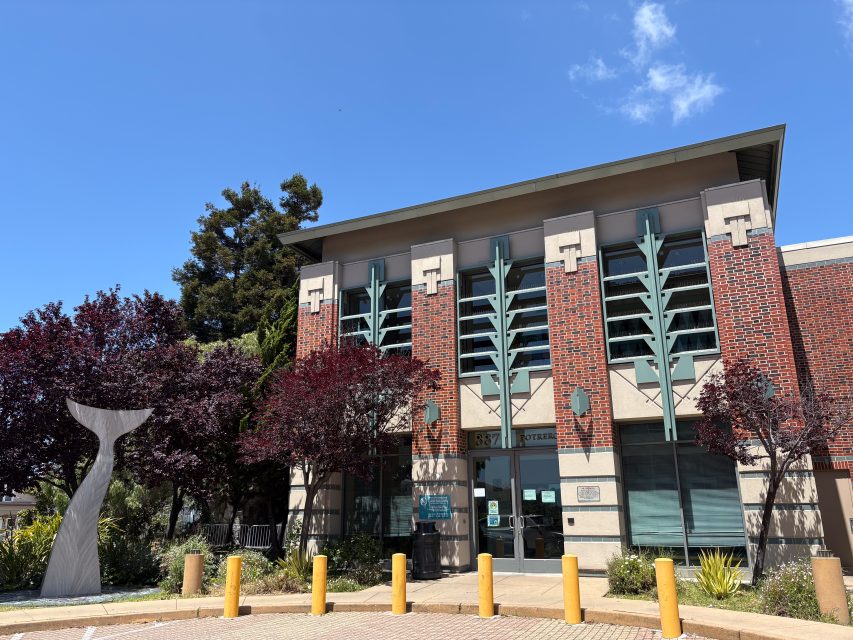 A two-story brick and concrete building with large windows and teal accents, featuring a whale tail sculpture and yellow bollards in front.