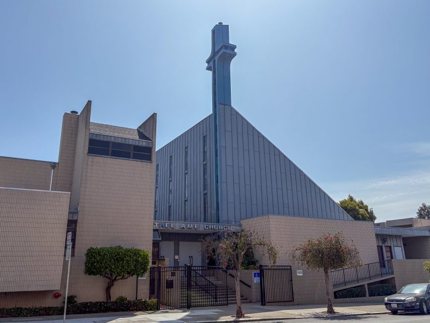 Modern church building with a large blue cross, beige brick exterior, and gated entrance on a sunny day.