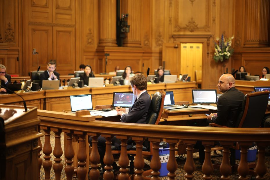 People seated at desks with laptops in a formal wood-paneled meeting room, possibly during a city council or government session.