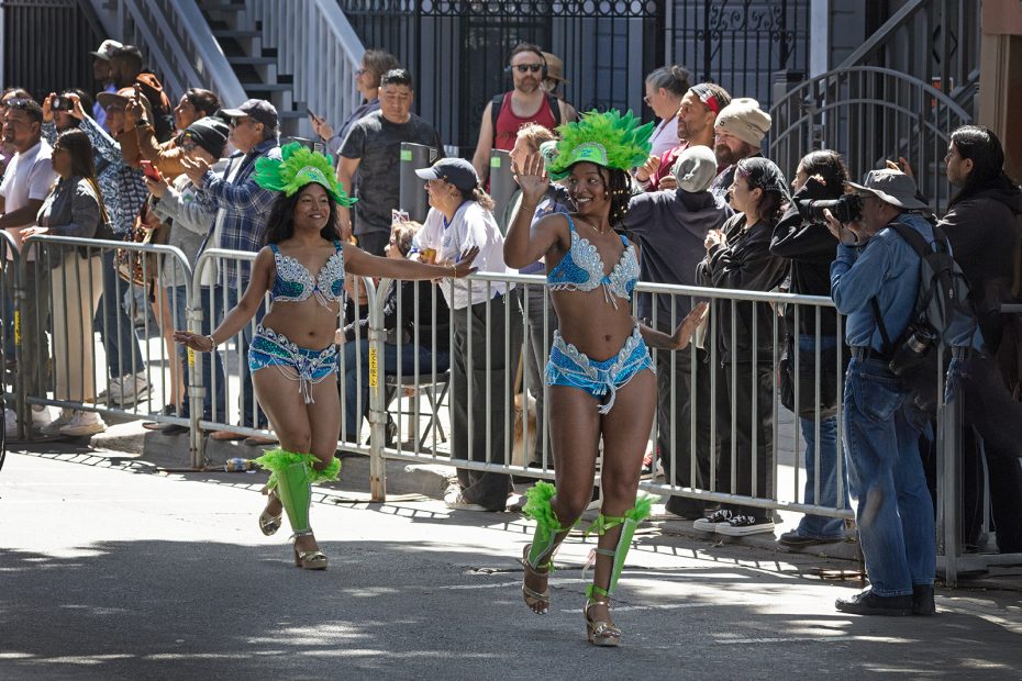 Two women in bright green and blue carnival costumes dance in a parade while spectators stand behind metal barriers, watching and taking photos.