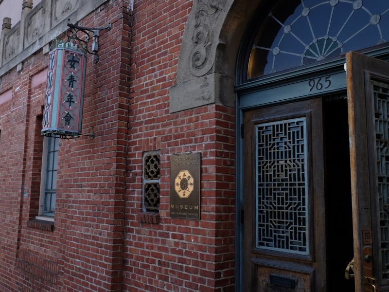 A brick building with a wooden door, a plaque reading "Chinese American Historical Museum," and a vertical sign with Chinese characters. Address number 965 is above the entrance.