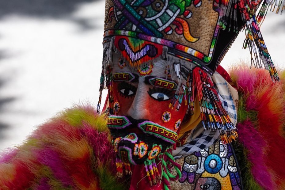 Person wearing a colorful, ornate traditional mask and headdress with intricate patterns and beads, and a vibrant fur garment.