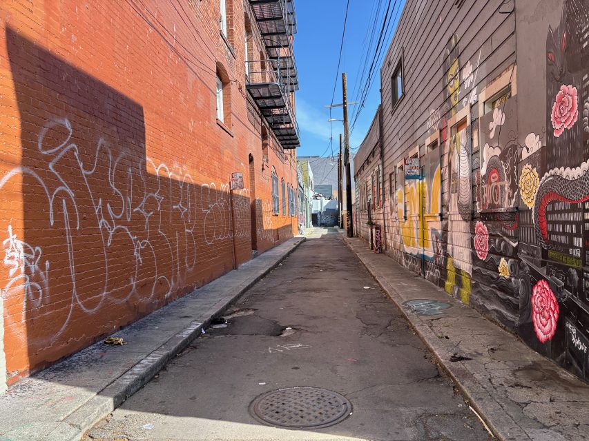 A narrow urban alleyway with graffiti on a red brick wall to the left and colorful murals on the right, under a clear blue sky.