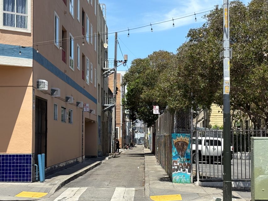 A narrow urban alleyway with buildings on the left, a fence and parked car on the right, string lights overhead, and some graffiti visible on the fence.