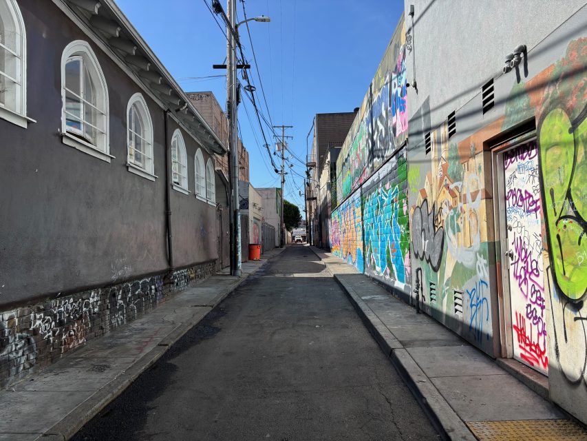 A narrow urban alleyway with one wall covered in colorful graffiti and the other painted gray, under a clear blue sky.