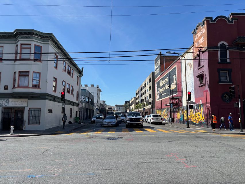 A city street intersection with cars waiting at a red light, pedestrians on sidewalks, and buildings with murals under a clear blue sky.