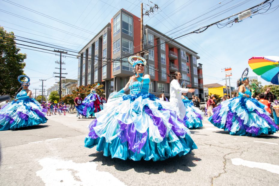 People in elaborate blue and purple dresses participate in a street parade on a sunny day, with a modern apartment building and electrical wires in the background.