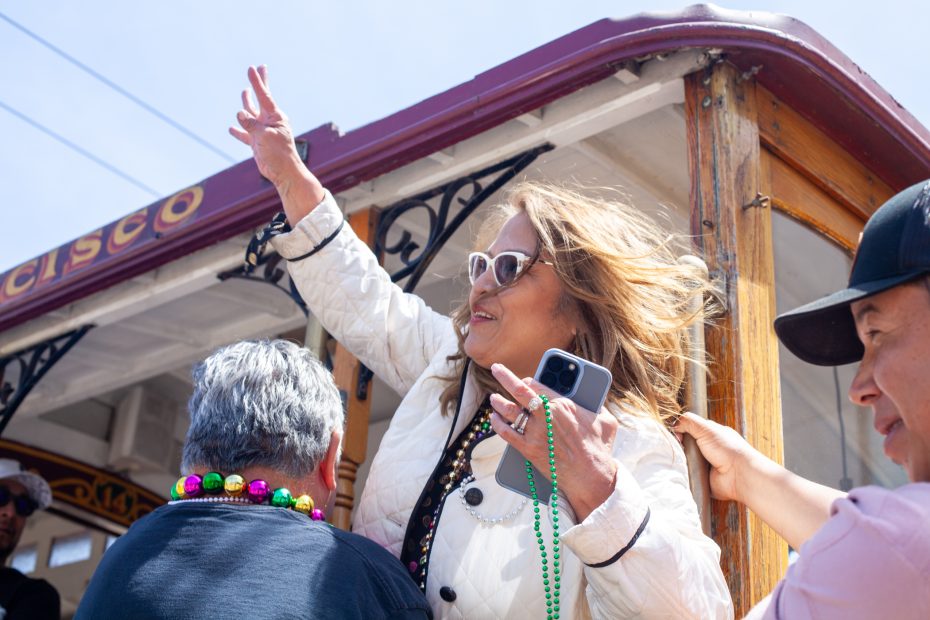 A woman wearing sunglasses waves while holding a phone and colorful beads on a San Francisco cable car, surrounded by other people.