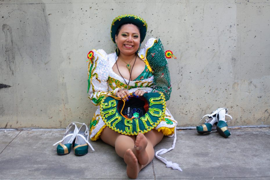 A woman in a colorful, traditional costume sits barefoot on the ground, smiling, with her shoes placed beside her and a decorative hat reading "San Simon" in her lap.