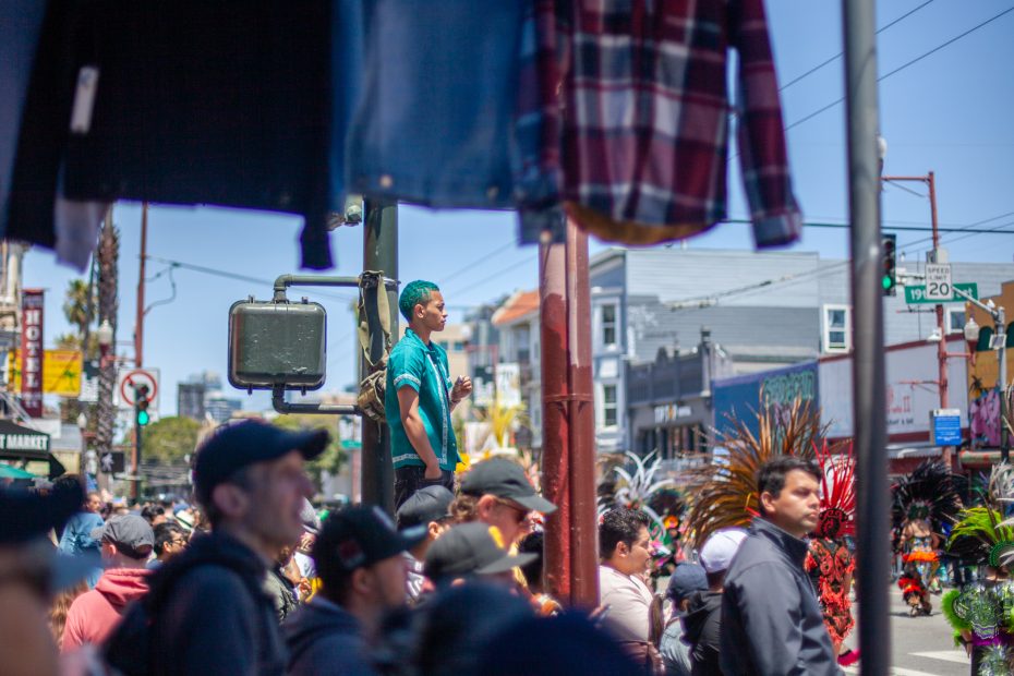 A crowd gathers at a busy street intersection during a parade; a boy stands on a pole for a better view, and feathered costumes are visible among the people.