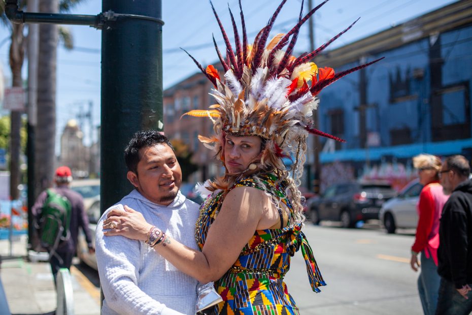 A person in a colorful, feathered headdress and patterned outfit poses with another person on a city street with people and cars in the background.