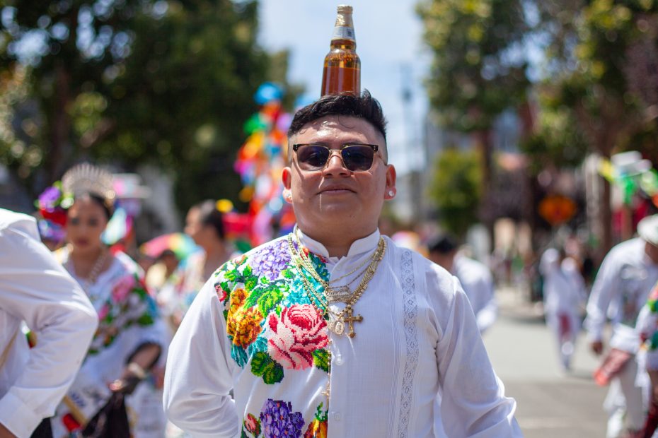 A man in traditional embroidered clothing balances a bottle on his head during a street parade, with other participants and colorful decorations in the background.