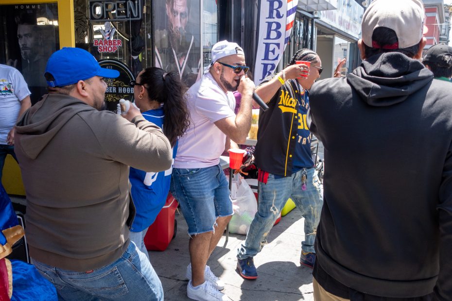 A group of people gather and dance on a city sidewalk near a barber shop during the daytime.