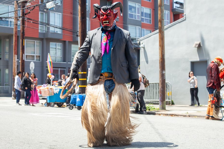 A person in a colorful demon mask, suit jacket, and long furry pants stands on a city street during a festival or parade.