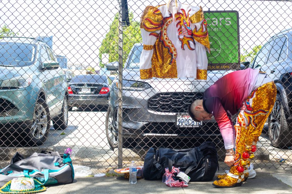 A person wearing a sequined costume bends down next to a chain-link fence in a parking lot, with a matching jacket hanging on the fence and bags on the ground.