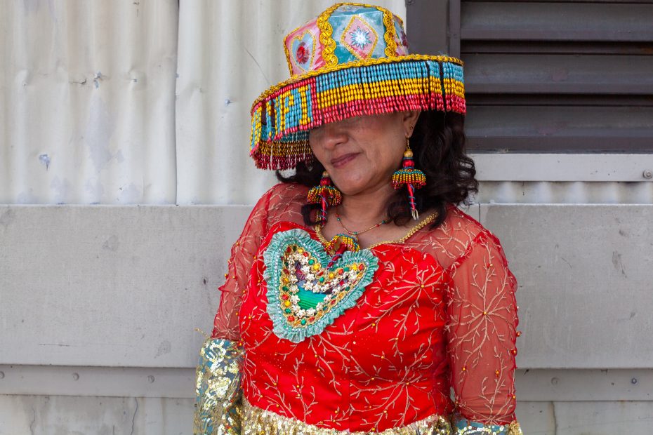 A woman in a colorful, beaded outfit and hat stands against a wall; her face is partially obscured by the hat’s beaded fringe.