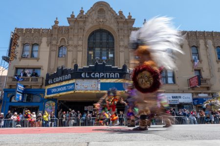 Dancers in colorful costumes move past El Capitan theater, with spectators watching from behind barriers on a sunny day.