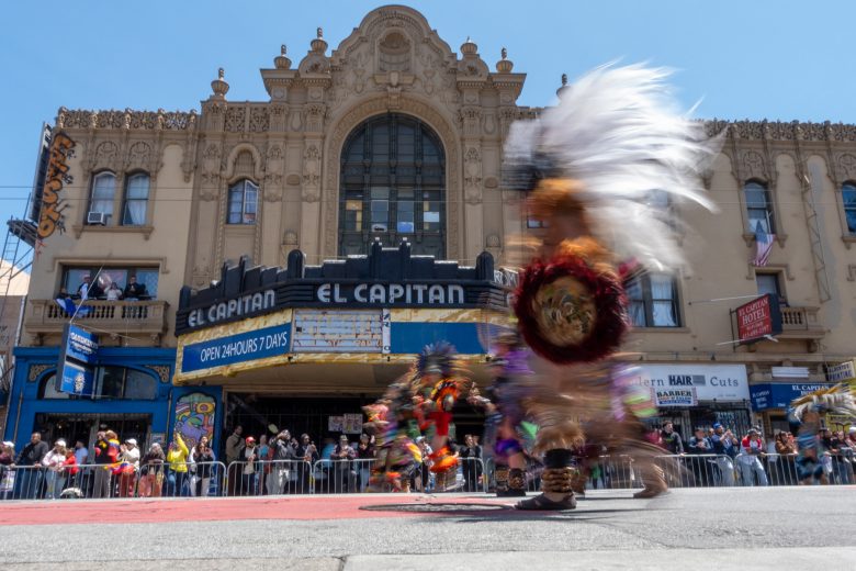 Dancers in colorful costumes move past El Capitan theater, with spectators watching from behind barriers on a sunny day.