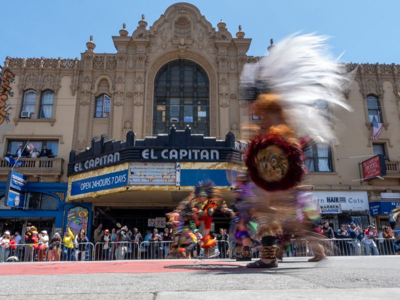 Dancers in colorful costumes move past El Capitan theater, with spectators watching from behind barriers on a sunny day.