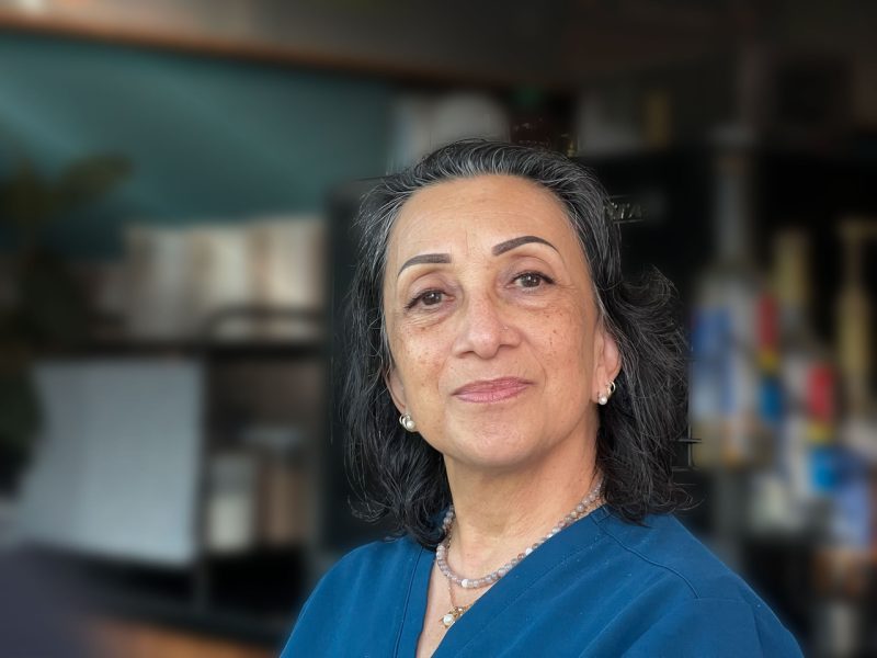 A middle-aged healthcare worker with gray hair and wearing a blue top looks at the camera with a neutral expression. The background is an indoor setting, slightly blurred.