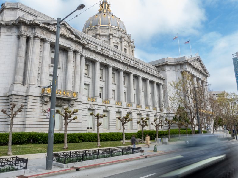 A grand government building with columns and a large dome, seen from the street with blurred motion of a passing car in the foreground.