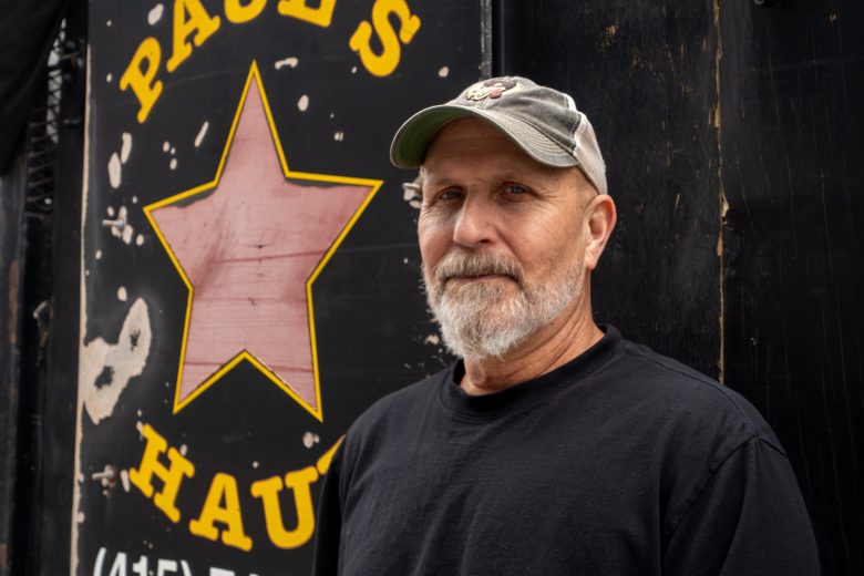 A man with a gray beard and cap stands in front of a black sign with a red star and yellow lettering.