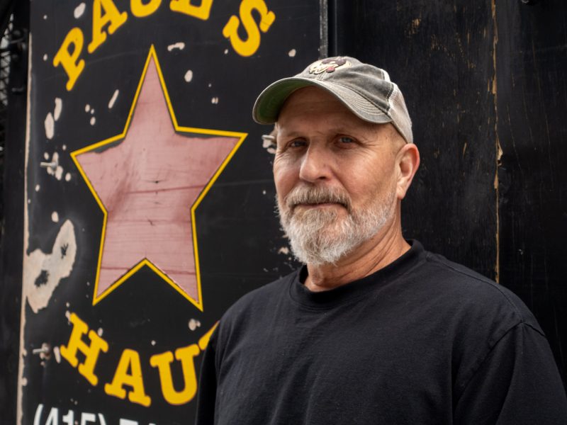 A man with a gray beard and cap stands in front of a black sign with a red star and yellow lettering.