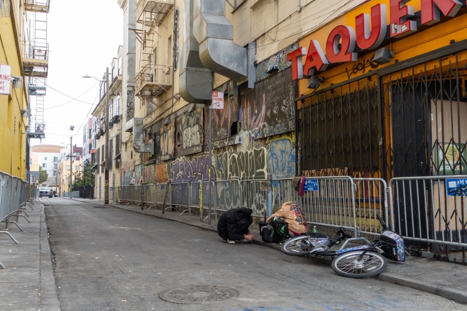 A person kneels beside another person lying on the ground next to a bicycle in a graffiti-covered alleyway near a closed taqueria.