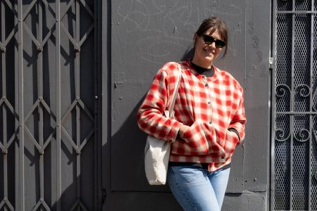 A person wearing sunglasses, a red plaid jacket, and blue jeans stands against a gray wall, holding a light-colored tote bag.