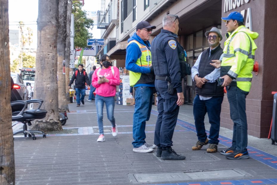 Several people, including a police officer and workers in safety vests, stand and talk on a city sidewalk while others walk by.