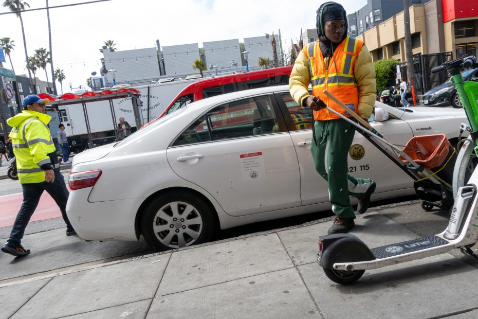 A worker in a safety vest moves a Lime scooter past a white city vehicle parked at the curb; another person in a yellow jacket walks nearby.
