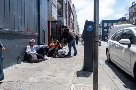 A person hands items to three people sitting on a sidewalk beside a building; a parking meter and parked cars are visible on the street.