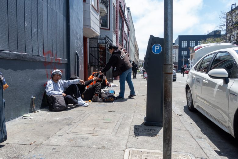 A person hands items to three people sitting on a sidewalk beside a building; a parking meter and parked cars are visible on the street.
