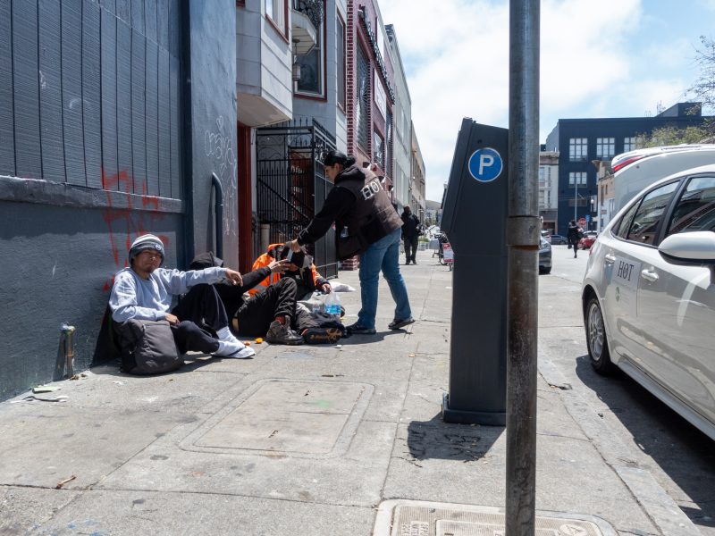 A person hands items to three people sitting on a sidewalk beside a building; a parking meter and parked cars are visible on the street.