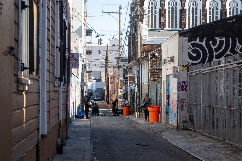 A narrow urban alley with graffiti, utility wires, and several people gathering near large orange trash bins in the background.