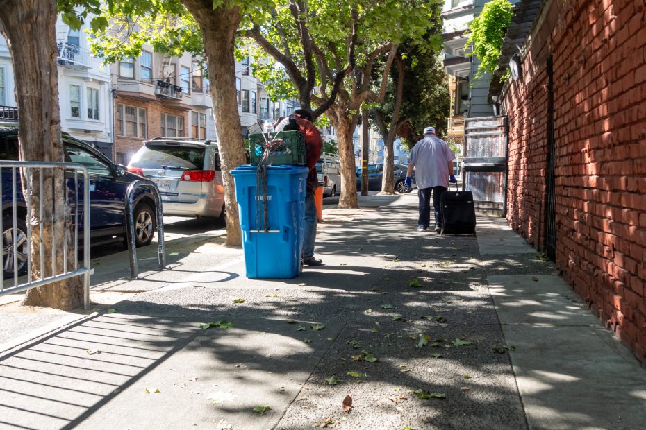 Two people on a city sidewalk, one handling blue recycling bins near parked cars and trees, while the other walks away pulling a black suitcase.