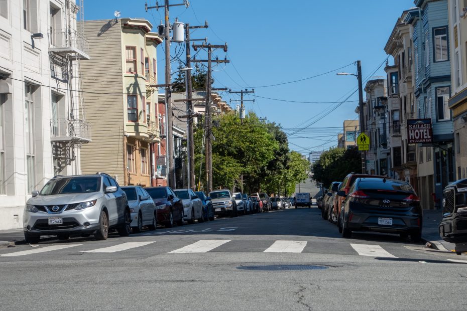 A city street with parked cars lining both sides, apartment buildings, utility poles, a crosswalk, and a clear blue sky.