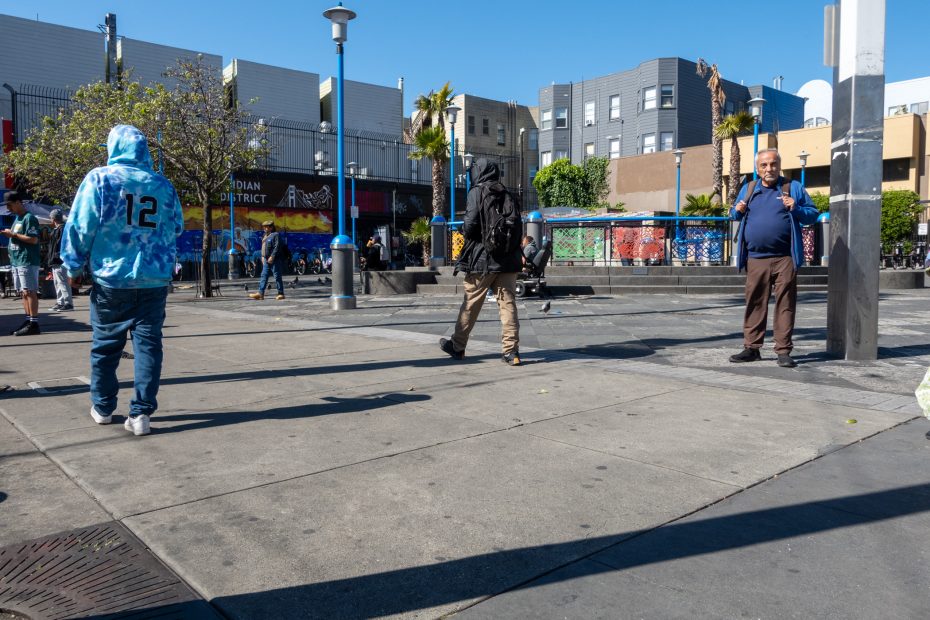 People walking and standing in an urban plaza on a sunny day, with buildings, trees, and colorful murals in the background.