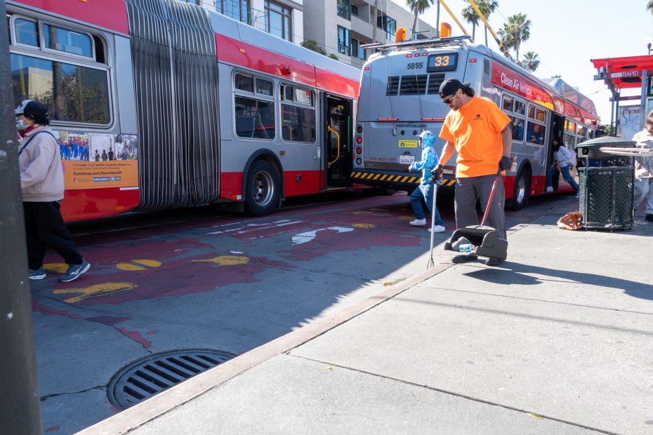 A person in an orange shirt sweeps the sidewalk near two city buses stopped on a street. Other pedestrians are visible nearby.